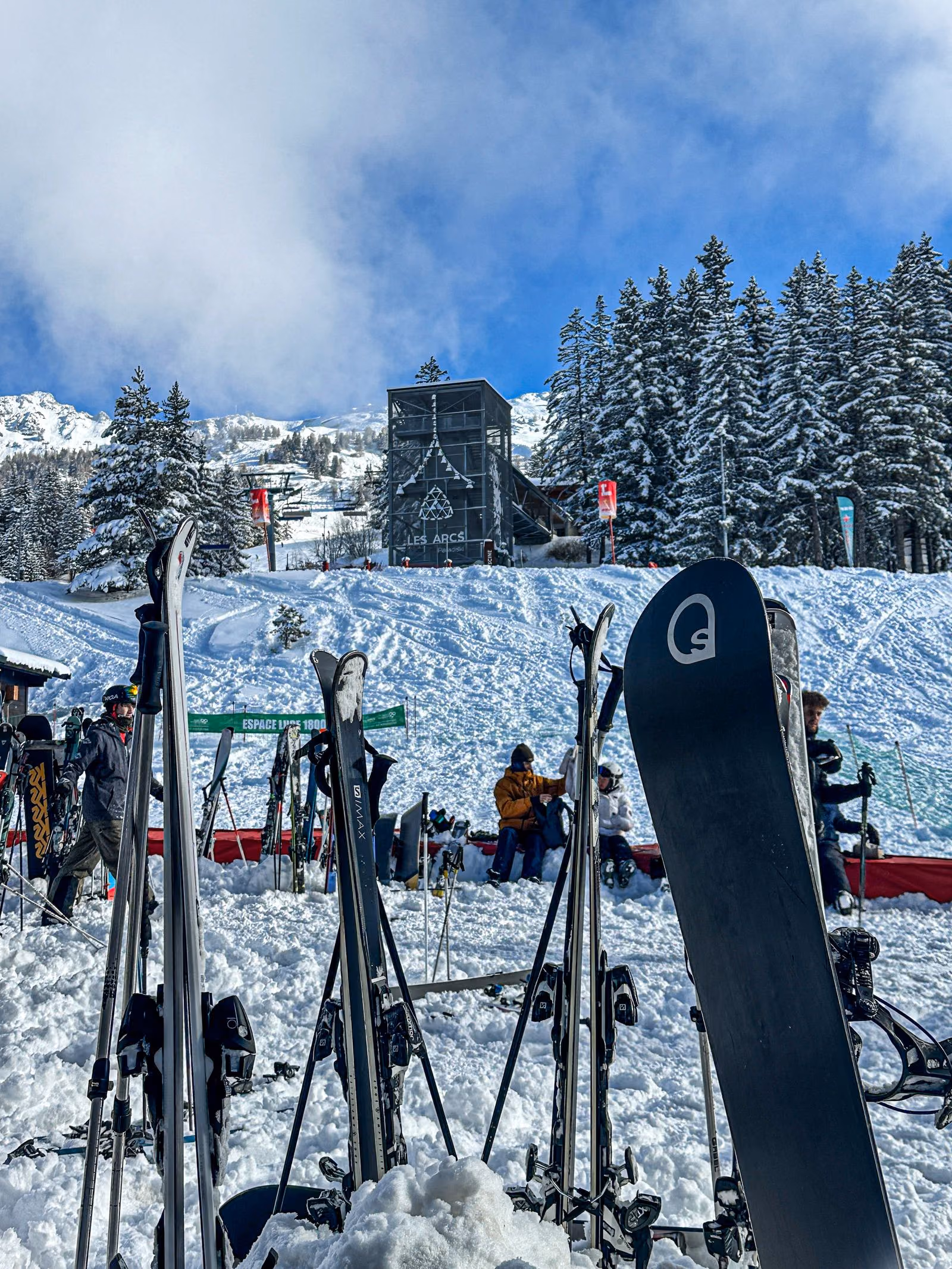 Des skis plantés dans la neige devant la gare de départ des Arcs 1800, par une journée d’hiver claire.