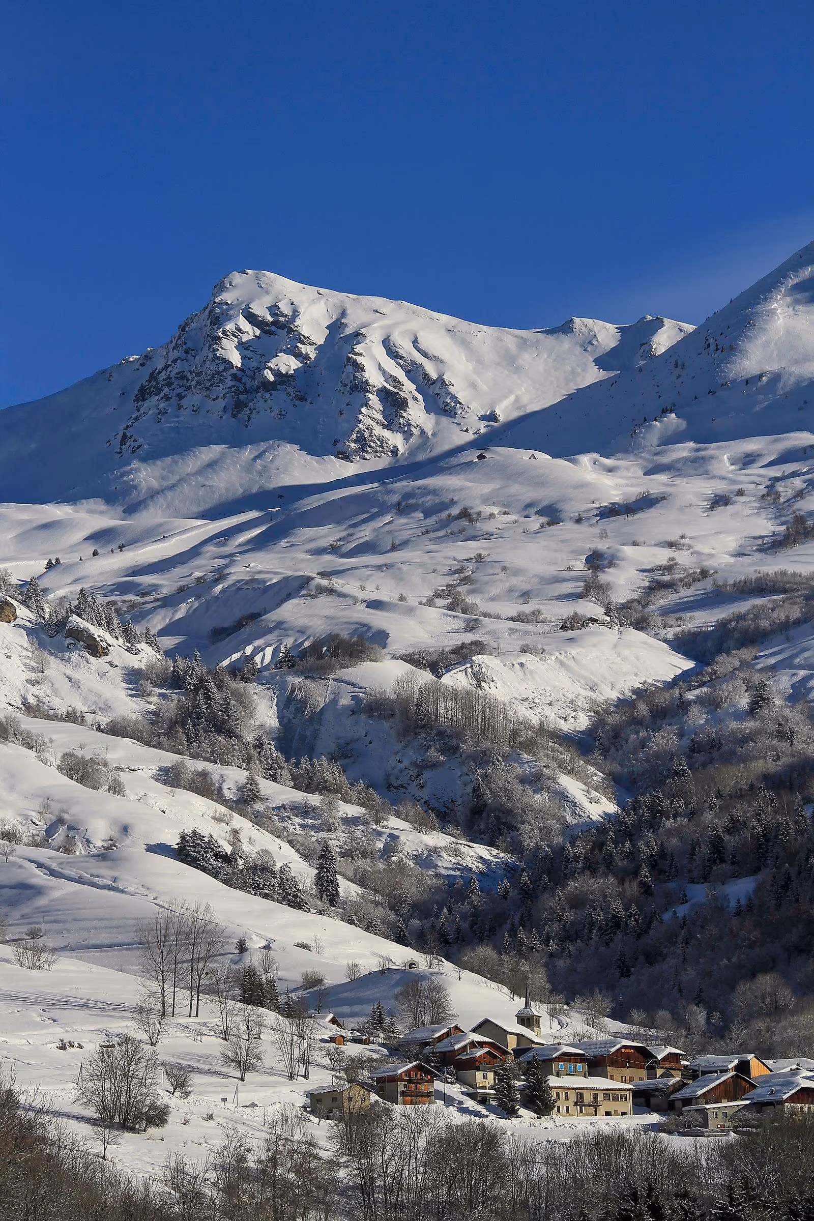 Un petit village savoyard et son clocher, dans un large vallon enneigé sous des pentes ouvertes.